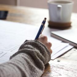 A person writing in a book at a table with coffee and papers